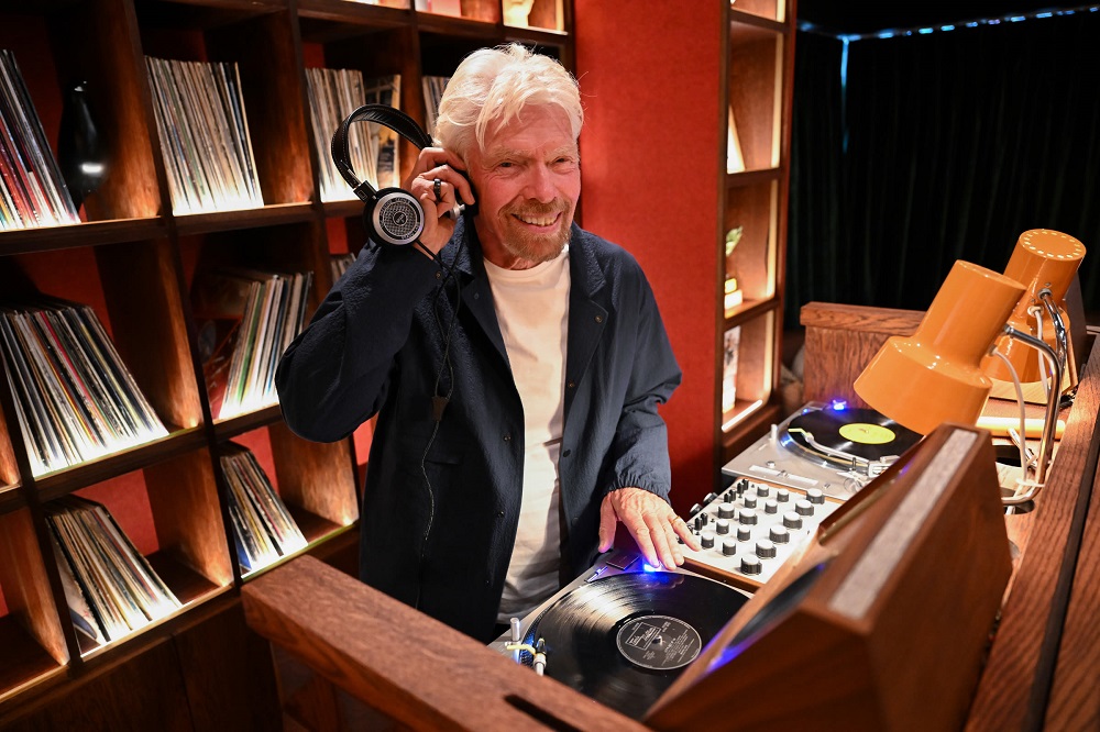 Dryly smiling DJ wearing headphones, operating turntables in a vinyl-club setup with shelves of records behind him.